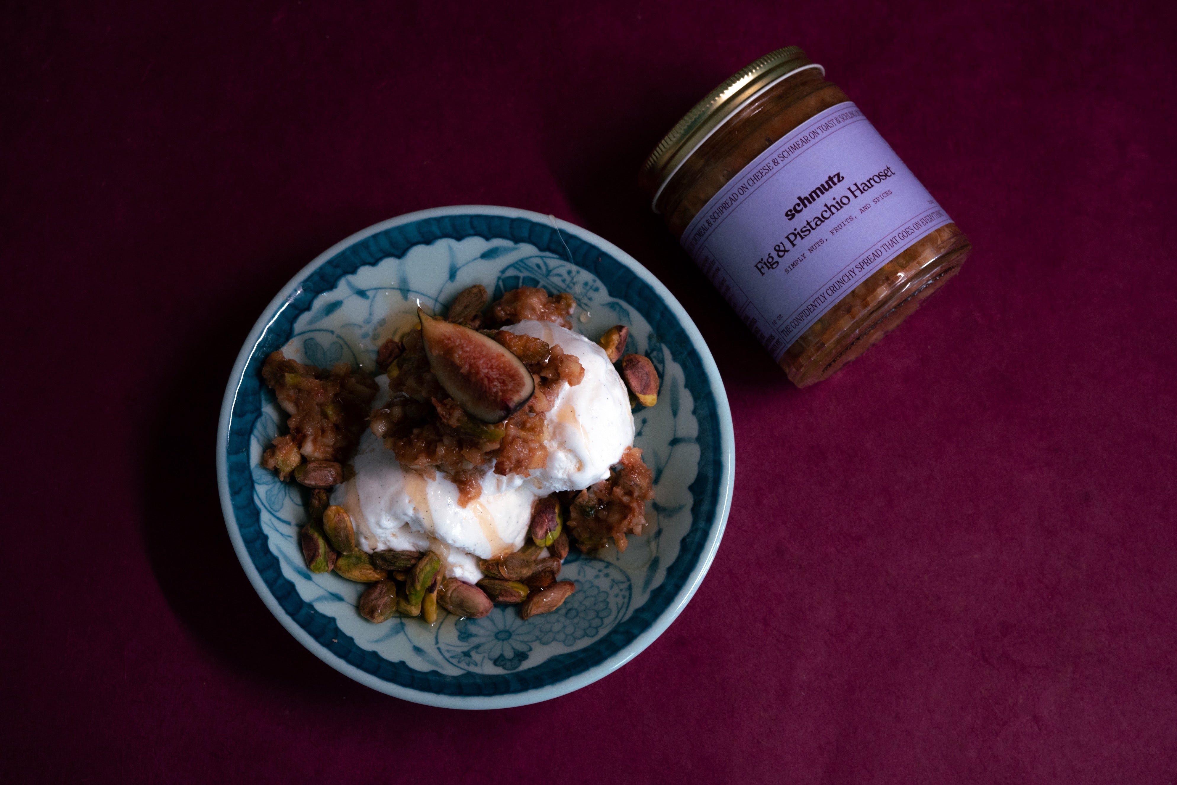 A bowl of vanilla ice cream topped with Schmutz Fig & Pistachio Haroset, next to a jar of Schmutz Fig & Pistachio Haroset, all on a deep purple background. Fig & Pistachio Haroset